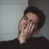 man wearing a brown jumper, holding his hand on his jaw due to sensitive teeth pain