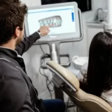Dentist wearing dark grey uniform, pointing at the screen showing Dentures, and patient sitting on a light brown chair
