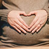 Pregnant woman wearing a brown jumper, with her hands on her belly shaping a heart